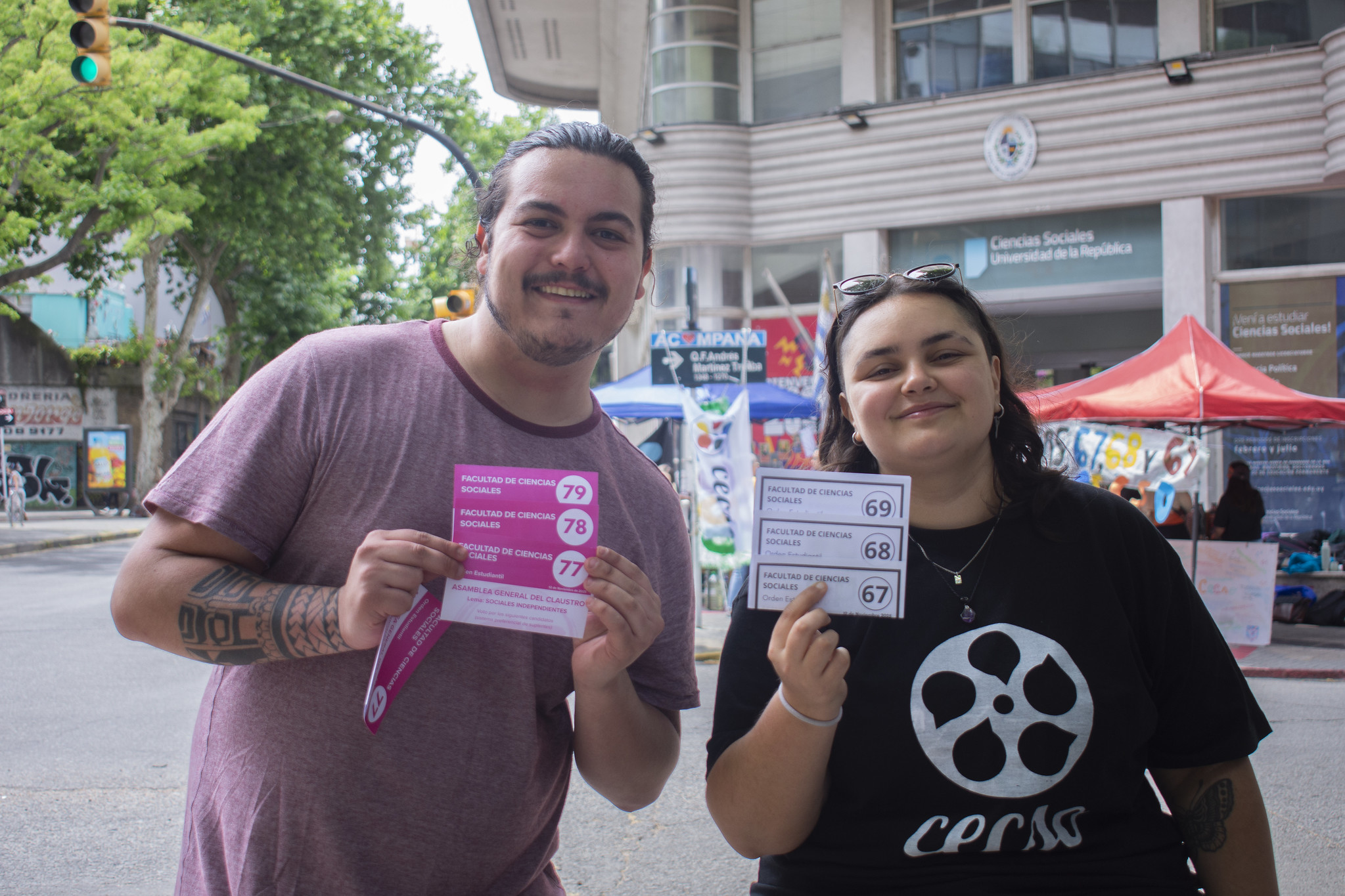 Un representante de Sociales Independientes y una representante del Centro de Estudiantes de Ciencias Sociales posan juntos mostrando sus listas en la puerta de la facultad.